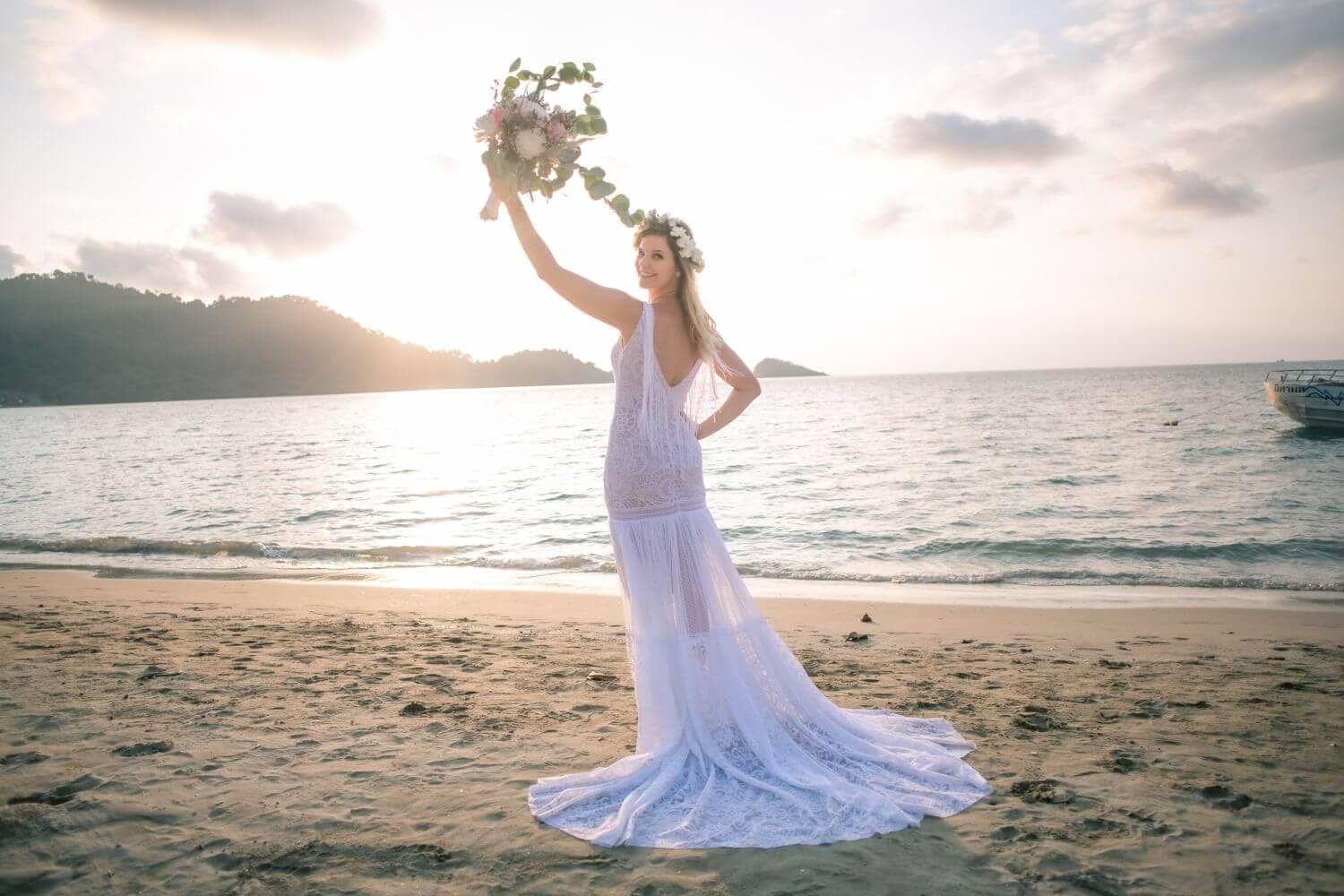Kobieta w białej sukni ślubnej trzymająca bukiet na plaży przy zachodzie słońca.