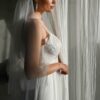 suknia ślubna z koronkowym gorsetem i długim welonem, widok z boku na kobietę stojącą przy zasłonie
